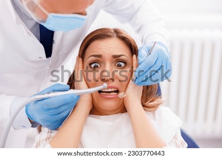 Woman afraid while sitting at dental chair at dentist office while doctor is holding dental drill and angled mirror, fixing patient's tooth
