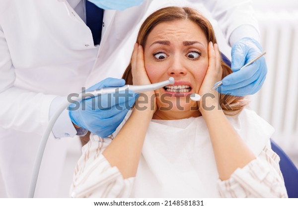Woman afraid while sitting at dental chair at\
dentist office while doctor is holding dental drill and angled\
mirror, fixing patient\'s\
tooth