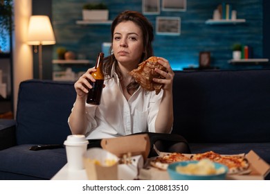 Woman after office work eating tasty takeaway hamburger holding beer bottle on couch watching news on television. Serious person having takeout burger and fast food delivery menu.