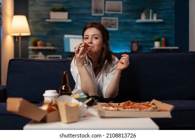 Woman After Office Work Taking A Bite Out Of Slice Of Hot Delivery Pizza While Watching Television At Table With Fast Food Takeway Menu. Person Sitting On Couch In Living Room Eating Takeout Dinner.