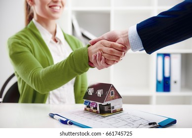 Woman agent shaking hands with new apartment owner over house model at the desk. 