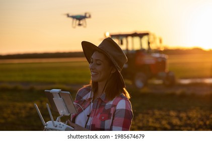 Woman Agronomist Using Drone Modern Technology In Agriculture. Young Farmer Standing In Field At Sunset In Front Of Tractor Machinery.