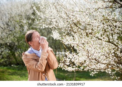 Woman allergic suffering from seasonal allergy at spring, posing in blossoming garden at springtime. Young woman sneezing in front of blooming tree. Spring allergy concept