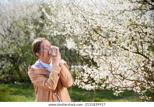 Woman allergic\
suffering from seasonal allergy at spring, posing in blossoming\
garden at springtime. Young woman sneezing in front of blooming\
tree. Spring allergy\
concept
