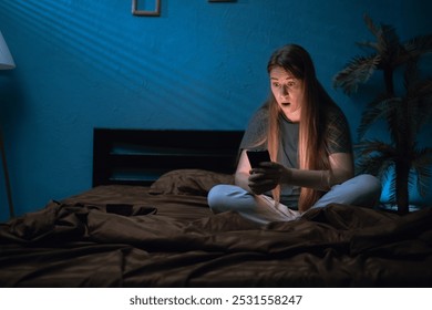 Woman is amazed by content reads shocking message on mobile phone sitting on her bed at night. - Powered by Shutterstock