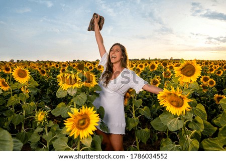 Woman among sunflowers wearing white dress and lifting hat smiling at sunset