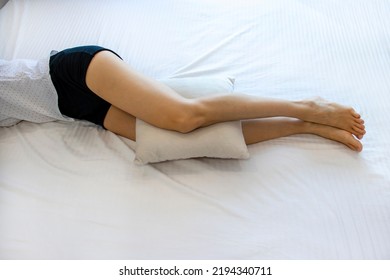 A woman with an anatomical pillow between her legs and knees, lying on a bed with white sheets. A girl resting on her side