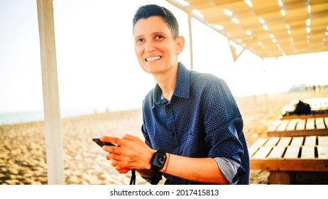 Eine Frau von androgynem Aussehen mit kurzen roten Haaren und einer Armbanduhr auf der Hand lächelt und hält ein Telefon auf einem Sandstrand. Hochwertiges Foto