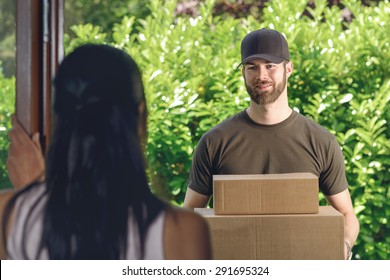 Woman Answering The Door To An Attractive Bearded Deliveryman Carrying Two Cardboard Cartons For Delivery, View Over Her Shoulder From Behind