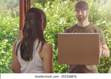 Woman Answering The Door To An Attractive Bearded Deliveryman Carrying Two Cardboard Cartons For Delivery, View Over Her Shoulder From Behind