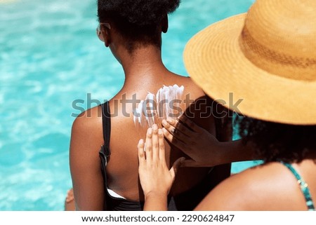 Woman applies sunscreen onto friend's back, sitting by pool wearing hat
