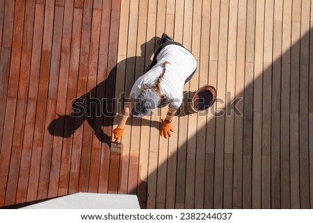 Woman applying brown wood protection oil on decking boards with paint brush, terrace renovation top view