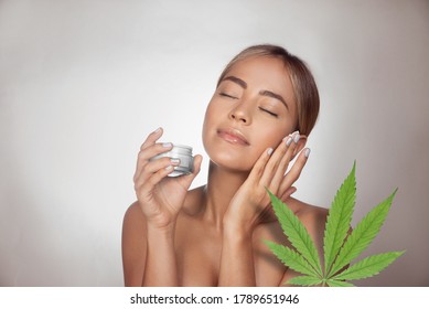 woman applying CBD facial cream made from cannabis extract for a natural skin treatment. Portrait of young woman with cannabis leaf. Cosmetology and treatment concept. Isolated on gray background