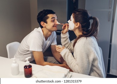 Woman Applying Clay Mask On Her Boyfriend's Face. Young Loving Couple Taking Care Of Skin At Home