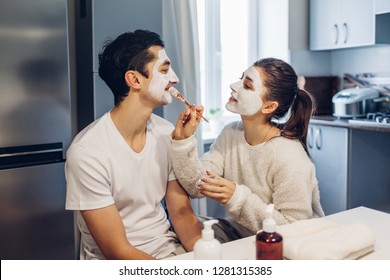 Woman Applying Clay Mask On Her Boyfriend's Face. Young Loving Couple Taking Care Of Skin At Home