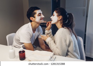 Woman Applying Clay Mask On Her Boyfriend's Face. Young Loving Couple Taking Care Of Skin At Home