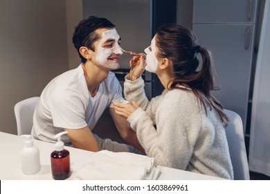 Woman Applying Clay Mask On Her Husband's Face. Young Loving Couple Taking Care Of Skin At Home. Healthy Routine