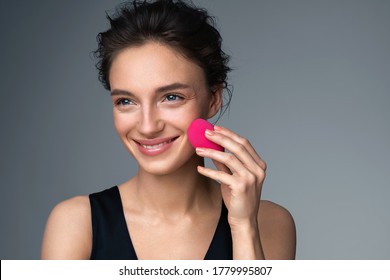 Woman applying foundation using cosmetic sponge, beauty blender. Photo of woman with perfect makeup on gray background. Beauty concept