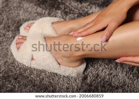 Woman applying health cream on legs. Girl wears modern puffy white slippers at home on carpet. Skincare and beauty concept