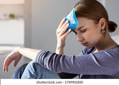 Woman Applying Ice Pack As Cold Compress On Forehead Due To Headache, Migraine, Tired After Work, Sitting Indoors In Apartment. Put Cold On Head For Relief Hurt And Fever