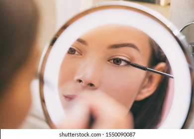 Woman applying make up beauty product putting mascara in ring lighted round makeup mirror at home bathroom morning routine. Beautiful Asian lady getting ready applying eye make-up with brush.
