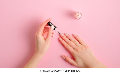 Woman Applying Polish On Nails At Home. Female Hands With Elegant Manicure And Nail Polish Bottle On Pink Background, Top View. Beauty Treatment And Hand Care Concept