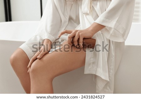 Woman applying self-tanning product onto her leg on tub in bathroom, closeup