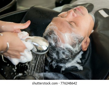 Woman Applying Shampoo And Massaging Hair Of A Customer. Men Which Wash Hair In A Beauty Salon. Hairdresser Washing Hair.Smiling Asian Caucasian Man Having His Hair Washed At The Hairdresser's,