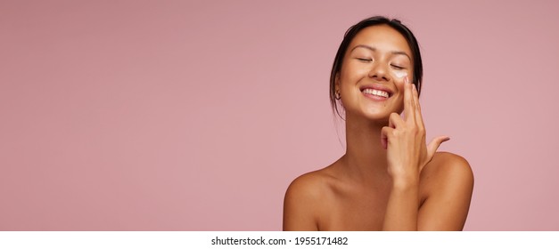 Woman applying skincare product on her face. Asian woman putting cosmetic cream in her face and smiling against pink background. - Shutterstock ID 1955171482