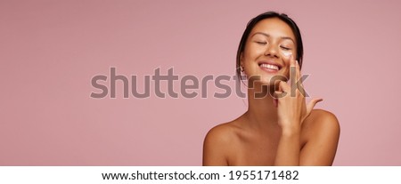 Woman applying skincare product on her face. Asian woman putting cosmetic cream in her face and smiling against pink background.