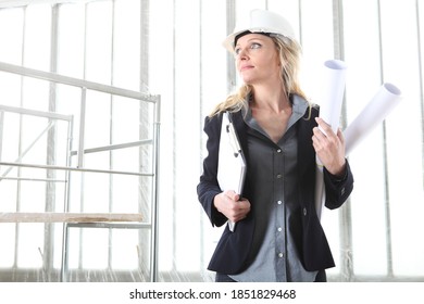 Woman Architect Or Construction Engineer Wear Helmet And Holds Folder And Blueprint Inside A Building Site With Windows And Scaffolding In The Background