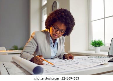 Woman architect drawing building plans on big format paper. Young African American lady sitting at her office desk with a laptop, holding a pencil, and working with modern house blueprint floorplans