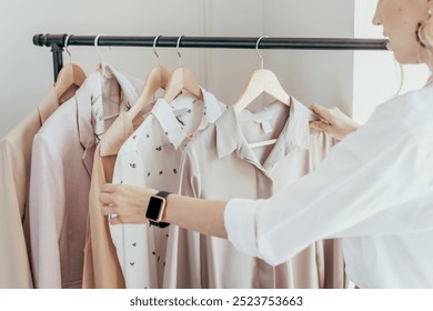 A woman arranges stylish fashion shirts on hangers. The woman is organizing shirts, focusing on white and beige shirts. The woman is wearing a smartwatch while handling fashion shirts. - Powered by Shutterstock