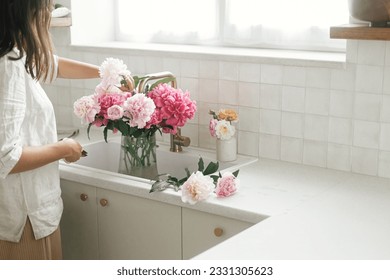 Woman arranging beautiful peonies in vase at sink with brass faucet in new modern kitchen. Female decorating flowers on background of granite countertop, hands with flowers close up - Powered by Shutterstock