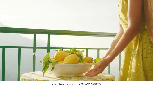 Woman arranging lemons on a balcony with an ocean view. Female adjusts a bowl of citrus fruits on table, sea backdrop glistening. Summer Italy. - Powered by Shutterstock