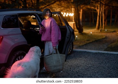 Woman arrives by car to a house in forest, standing with bag and phone near vehicle in the evening time. Traveling by car and rest in cabins on nature concept - Powered by Shutterstock