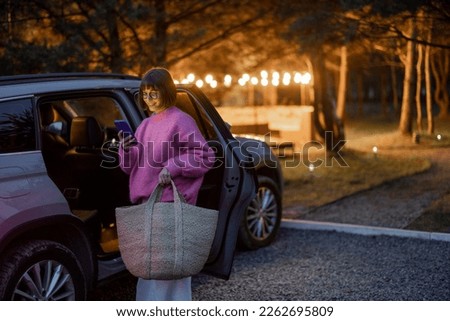 Woman arrives by car to a house in forest, standing with bag and phone near vehicle in the evening time. Traveling by car and rest in cabins on nature concept