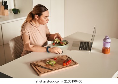 Woman With Artificial Limb Eating Sandwiches At The Table While Watching Something At The Laptop