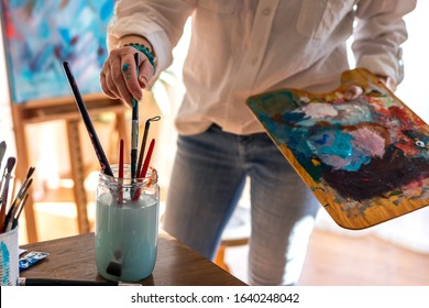 Woman artist cleaning paintbrush during working on her abstract paintings at art studio. Pallete with acrylic colors in female hand. Close-up selective focus - Powered by Shutterstock