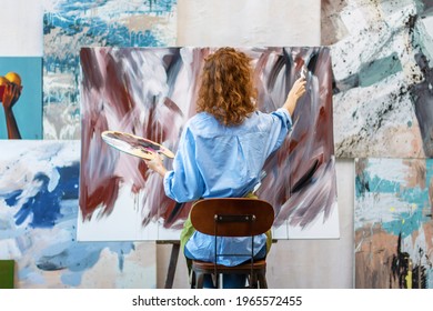 Woman artist is drawing on canvas with brush, palette, in workshop studio gallery. Process of making oil, watercolor, acrylic painting at workplace with tools, supplies for creation. Contemporary art. - Powered by Shutterstock