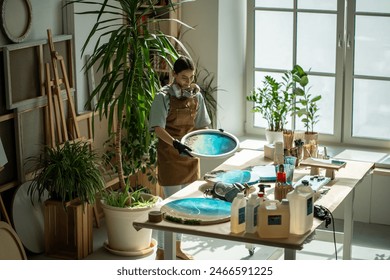 Woman artist examining finished acrylic sea painting on wooden circle canvas covered with epoxy resin. Painter work in art studio with indoor plants. Home art studio, small business, hobby, liquid art - Powered by Shutterstock