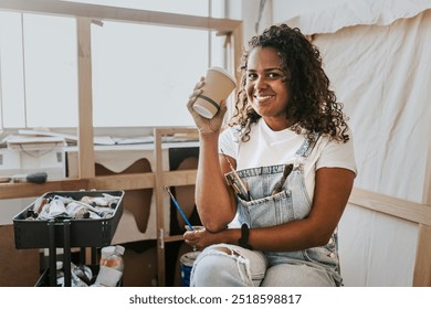 Woman artist holding a paper coffee cup, happy female artist in art studio with sketches and canvases, art and creativity - Powered by Shutterstock