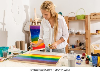 Woman artist painting lgbt flag while standing at table and paints in art studio adding orange color