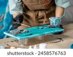 Woman artist pours epoxy resin and smears it with stick on wooden board. Craftswoman using liquid art technique, creating picture of sea foam and waves, rocks closeup view. Master class on resin art. 