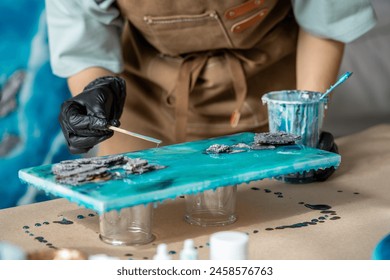 Woman artist pours epoxy resin and smears it with stick on wooden board. Craftswoman using liquid art technique, creating picture of sea foam and waves, rocks closeup view. Master class on resin art.  - Powered by Shutterstock