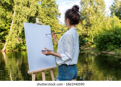 woman artist in summer park by lake, background forest water shore trees, draws a picture, view from back, in hand palette with paints, brush and easel. Jeans clothes white shirt. White blank canvas. - Powered by Shutterstock