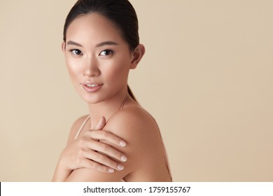 Woman. Asian Model Beauty Portrait. Beautiful Female Touches Her Shoulder And Looking At Camera. Tender Girl With Perfect Glowing Skin And Nude Natural Makeup Posing Against Beige Background. 