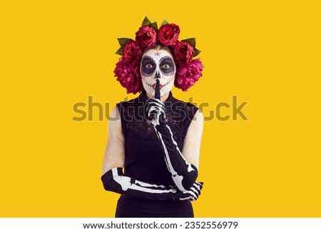 Woman asks to keep her secret on Day of Dead or Halloween. Lady in Catrina costume with sugar skull face and wreath of beautiful roses doing shhh tss sign standing isolated on color studio background
