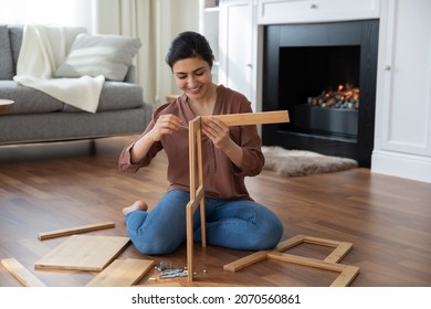 Woman assembling new furniture at home, new purchase, hobby concept. Young Indian female sit on warm floor in living room with fireplace holding wooden boards chair details, collects modern furniture