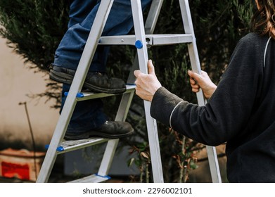 A woman assistant holds an aluminum stepladder, insuring a worker, a man, a builder in a dirty robe standing on the steps. Photography, portrait of legs, shoe close-up, lifestyle, outdoor work. - Powered by Shutterstock